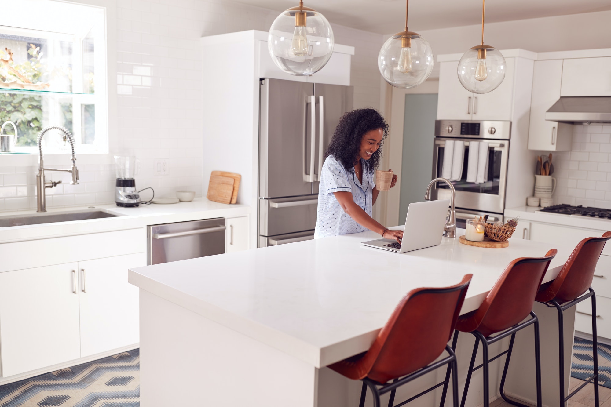 Woman Wearing Pyjamas Standing In Kitchen Working From Home On Laptop